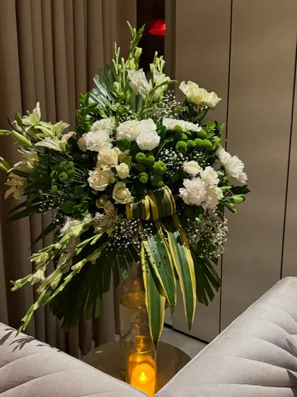 A classic arrangement of white lilies, carnations, and lush greenery in a glass vase, placed next to a sofa. A simple touch that instantly elevates the space.