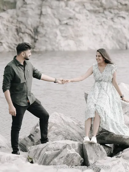 A couple holds hands while standing on rocks by a lake, creating a romantic and scenic pre-wedding portrait.