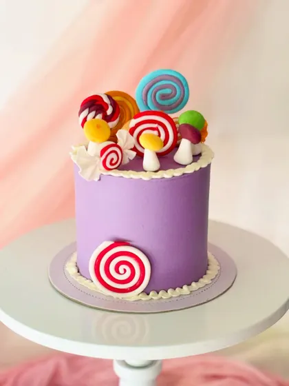 A vibrant purple cake covered in colourful, oversized fondant lollipops and candies. The simple piped border adds a classic finishing touch.