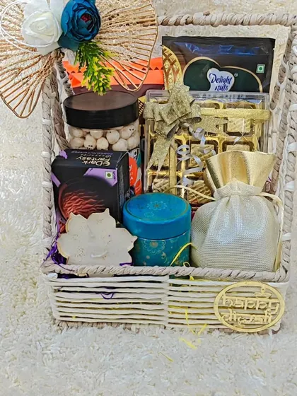 A close-up of a white wicker basket hamper filled with a delightful mix of treats. It includes roasted makhana, cookies, Omani dates, a decorative candle, and a potli bag of chocolates.