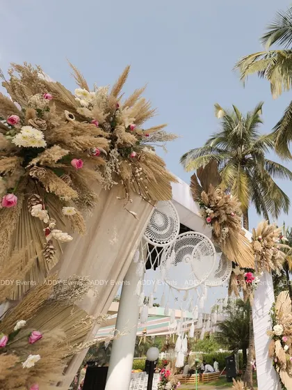 A low-angle view of our signature boho wedding arch, highlighting the rich textures of pampas grass, dried spear palms, and delicate roses against a clear blue sky. The addition of macrame dreamcatchers adds a whimsical touch.