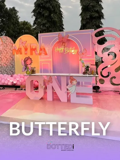 An enchanting butterfly-themed first birthday for Myra. The outdoor setup features pastel-colored backdrops, a "Happy Birthday" neon sign, and large butterfly cutouts.