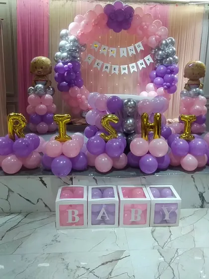 A full stage setup for the naming ceremony of 'Trishika', with a pink and purple balloon ring, 'BABY' blocks, and the name in gold foil letters.