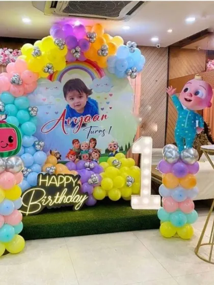A bright and cheerful Cocomelon theme for a first birthday party. This setup includes a custom backdrop with the birthday boy's photo, colorful balloon arrangements, character cutouts, a 'Happy Birthday' neon sign, and a light-up number '1'.