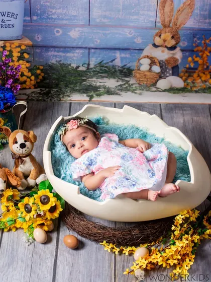 A happy baby girl lies in an eggshell prop on a bed of soft blue fabric. The scene is decorated with sunflowers, stuffed animals, and Easter eggs, creating a cheerful spring-themed infant photo.