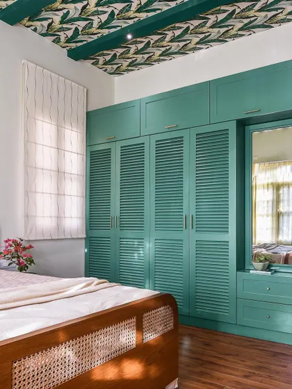 This bedroom features a stunning teal wardrobe with louvered doors and an integrated dressing mirror. The design is paired with a leaf-patterned wallpaper on the ceiling for a touch of tropical drama.