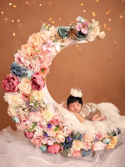 A newborn baby wearing a tiny crown sleeps on a crescent moon prop lavishly decorated with pastel-colored flowers. This is a perfect example of a high-concept, fantasy-themed newborn shoot.