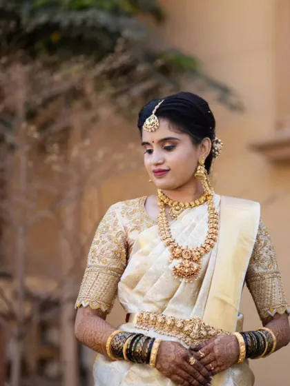An elegant muhurtham look for Divya. The classic combination of a white silk saree and temple jewellery was enhanced with subtle, defining makeup and a traditional hair bun.