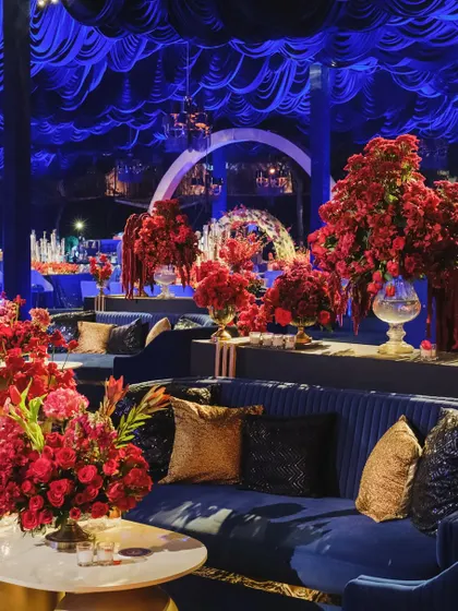 A wide shot of the moody blue and red Sangeet, showing the deep blue draped ceiling, velvet sofas, and the dramatic arches in the background.