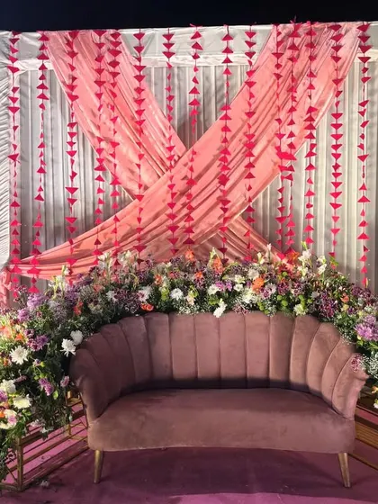 A romantic and pretty Mehendi seating arrangement. A dusty pink scalloped sofa is nestled in a bed of multi-coloured flowers, with draped curtains and hanging paper garlands creating a soft backdrop.