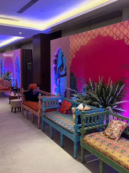 A luxurious Mehendi setup with traditional Indian seating (diwans) in vibrant colors. The backdrop features peacock motifs and intricate patterns, creating a royal ambiance.