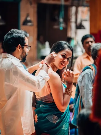 A playful moment during a pre-wedding shoot in a bustling market. The groom-to-be gently places flowers in his partner's hair, showcasing a sweet, unposed interaction.