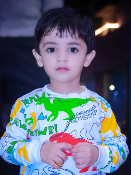 A close-up portrait of a young boy in a colorful dinosaur-themed shirt, looking directly at the camera.