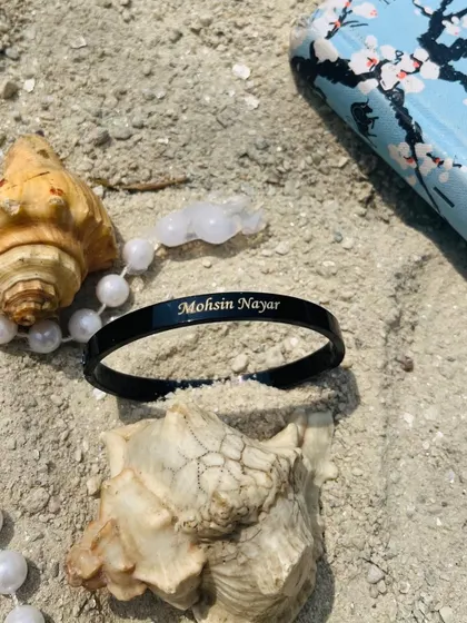 A sleek black Kada with the name 'Mohsin Nayar' engraved, styled on the sand with seashells. The black finish offers a modern and bold alternative.