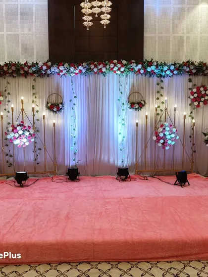 A simple yet elegant stage with a pink carpet and white drape backdrop. The hanging floral baskets and warm lighting add a touch of class, suitable for an engagement or a reception party.