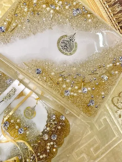 An Eid gift set featuring a Quran box and other accessories in a stunning white and gold design, embellished with pearls and crystals. It's laid out on a golden velvet prayer mat.