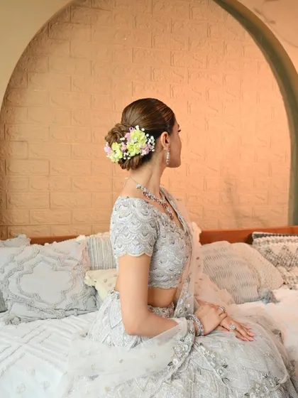 A beautiful shot of the bride seated, highlighting the elegant floral bun and the delicate details of her silver lehenga. The makeup is soft and luminous.