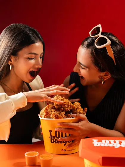 A lifestyle shot for Louis Burger showing two people excitedly digging into a bucket of fried chicken. This image captures the joy and social aspect of sharing food.