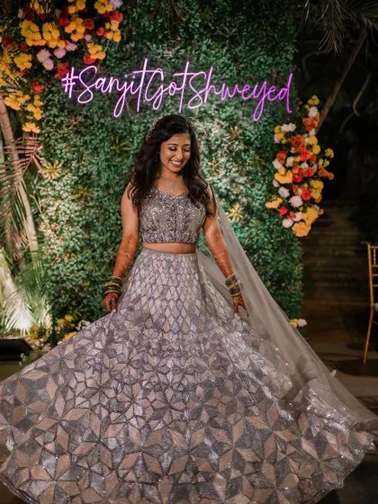 A candid moment of pure joy. The bride steals the spotlight at her sangeet night, posing in front of a personalized neon sign in her stunning silver lehenga.