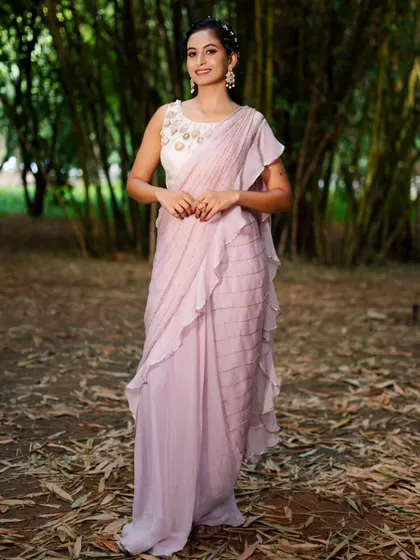 This lavender pre-draped saree features delicate ruffles and sequin work. It's paired with a sleeveless white blouse with intricate gold and pearl embroidery on the neckline.
