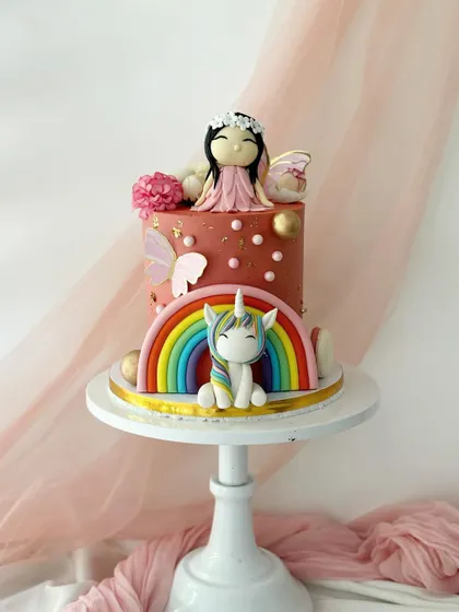 A whimsical cake featuring a fairy and a unicorn. The design includes a fondant rainbow, butterflies, and golden spheres for a touch of enchantment.