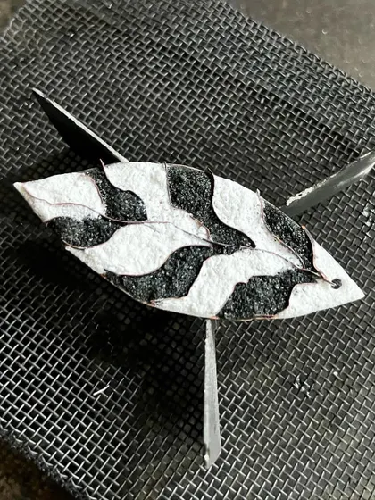 An enamel piece during the firing process, resting on a mesh before going into the kiln where the glass powder melts and fuses to the copper.