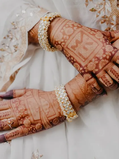 A detail shot of the bride's hands, adorned with intricate henna and beautiful white bangles, resting together in a moment of quiet anticipation.