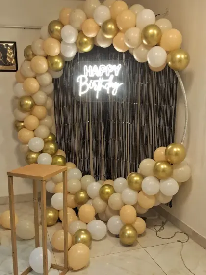 A chic and modern birthday backdrop with a circular frame, a garland of gold, cream, and white balloons, and a 'Happy Birthday' neon sign against a black shimmer curtain.