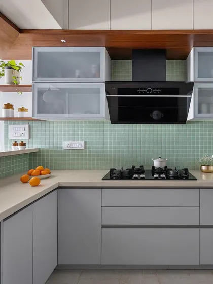 A close-up of the 'Gulmohar' kitchen, showing the soft grey-green tile backsplash, frosted glass cabinets, and warm wood details.