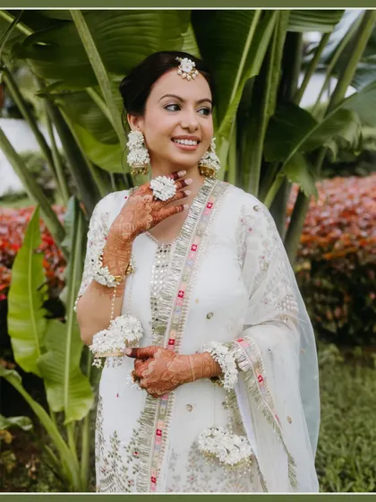 A smiling bride in a white and gold outfit adorned with floral jewelry stands against a lush green background, looking fresh and radiant.