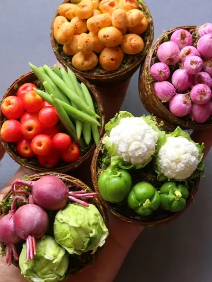 A hand holding five baskets of incredibly realistic miniature vegetables, including tomatoes, beans, and cauliflower.