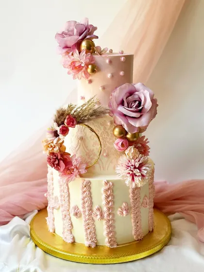 A breathtaking three-tier cake combining multiple textures and techniques. It features stenciling, knitted piping details, polka dots, and is adorned with a wreath of silk flowers and pampas grass.