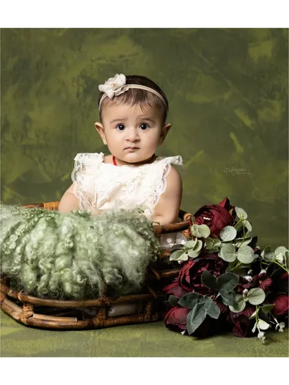 A 6-month-old baby, sitting up in a basket against a rich green backdrop. The deep red flowers add a beautiful pop of color.