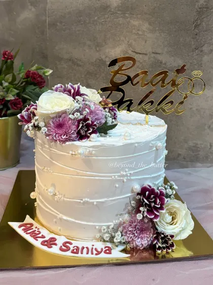 A beautiful cake for a beautiful "Baat Pakki" ceremony. This design features textured white buttercream, a cascade of fresh purple and white flowers, and a custom topper to celebrate the happy couple, Aijaz and Saniya.