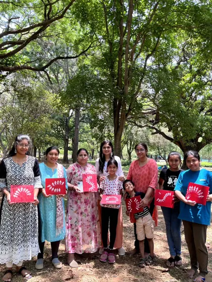 A group of women and children proudly hold up their completed Warli paintings on bright red canvases. It's rewarding to see a mix of ages enjoying and learning a traditional art form together.