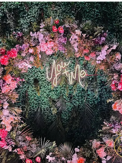 A "You + Me" neon sign set within a lush floral and green wreath, a perfect modern photo backdrop.