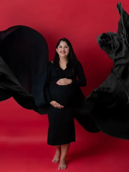 A powerful solo maternity portrait featuring a woman in a black gown against a vibrant red background. The gown's fabric is tossed to create a dynamic, wing-like shape, resulting in a bold and modern studio photograph.