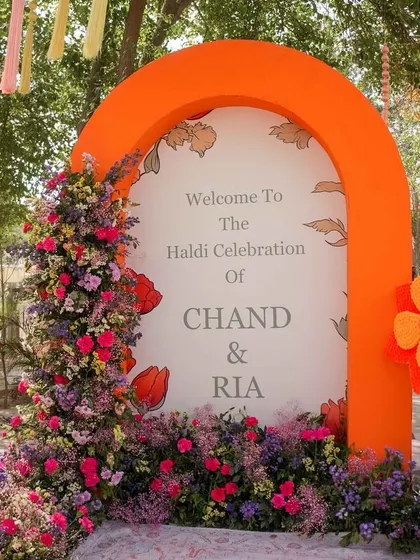 A bright and welcoming entrance for a Haldi celebration. The bold orange arch is adorned with a cascade of colorful fresh flowers, setting a cheerful tone for the event.