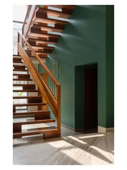 This wooden staircase in the 'Solid Void Solid' house is set against a deep green feature wall, with sunlight creating a dynamic play of shadows. It serves as a warm, tactile element that connects the different levels of the home.