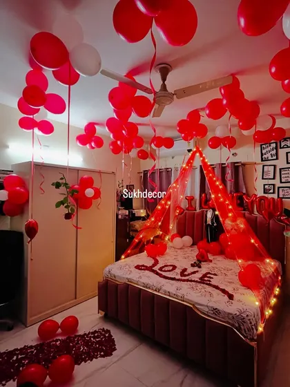 We can create a romantic canopy over the bed using sheer fabric and fairy lights. This setup features a ceiling full of red and white balloons, a "LOVE" message made of rose petals, and a beautiful lit canopy for a dreamy atmosphere.