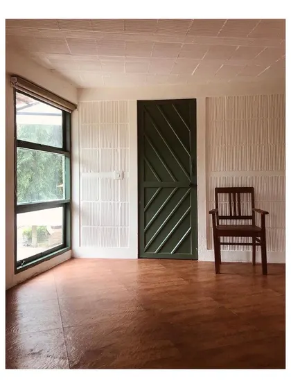 An empty room in a farmhouse project, highlighting the core architectural elements. The rich, earthy oxide flooring contrasts with the white textured walls and the dark green door, creating a simple yet characterful space.