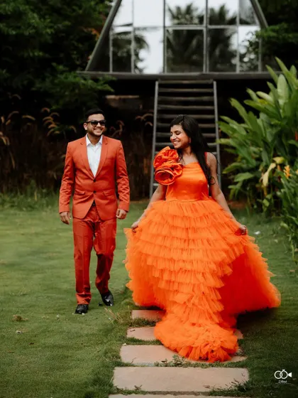 The couple walks through a garden, the bride's magnificent ruffled orange gown creating a stunning visual against the green lawn.