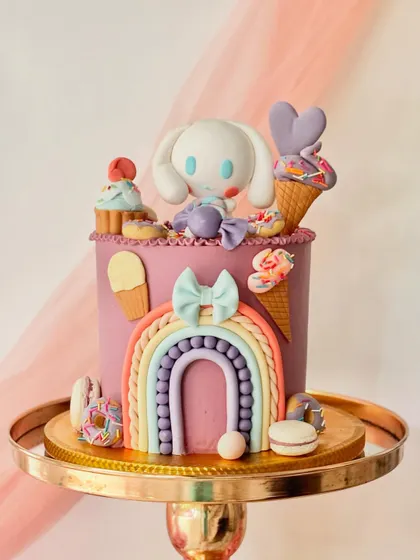 This cake is a sweet treat explosion, featuring the Sanrio character Cinnamoroll. It's surrounded by fondant rainbows, ice cream cones, donuts, and macarons in a pastel colour palette.