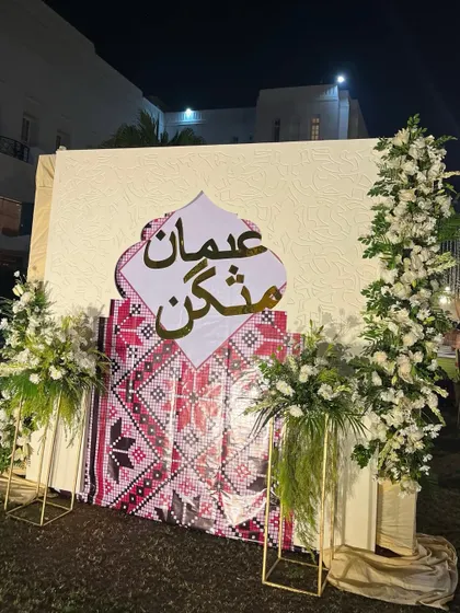 A tribute to tradition. This backdrop features elegant Arabic script and a vibrant patterned fabric, framed by pillars of white flowers, celebrating cultural heritage.
