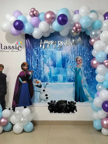 A magical Frozen theme party with a winter forest backdrop, life-size cutouts of Elsa, Anna, and Kristoff, and a balloon arch in icy blue, white, and purple.