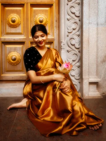 Seated by a traditional golden door, the lustrous fabric of this gold satin saree catches the light beautifully. Paired with a simple black velvet blouse, the look is one of pure elegance.