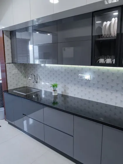 This kitchen features glossy grey handleless cabinets, a black granite countertop, and a 3D-textured backsplash. The glass-fronted upper cabinet with internal lighting is perfect for displaying crockery.