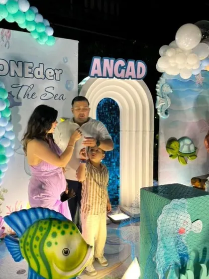 A family enjoys a moment at their son Angad's "ONEder The Sea" themed first birthday party, complete with a waterfall backdrop and aquatic props.