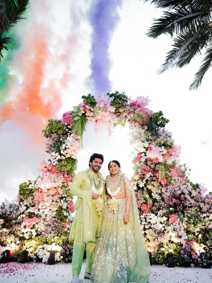 The couple stands under a magnificent floral arch as colorful smoke bombs create a rainbow in the sky behind them, a truly magical moment.