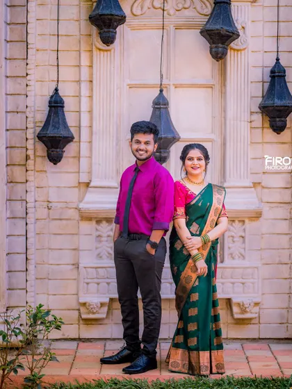 A classic portrait of a couple in traditional attire, set against a wall with hanging lanterns. The simple, elegant pose highlights their beautiful outfits.